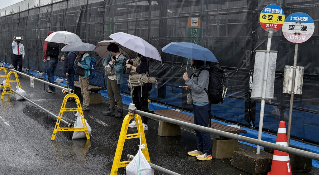 雨のなか傘を手に路線バスの到着をじっと待つ観光客ら=2025年11月5日、屋久島空港、屋久島ポスト撮影