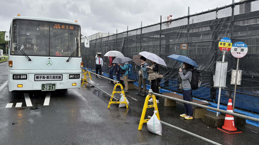 雨に打たれながら傘を手に路線バスを待つ観光客ら=2025年11月5日、屋久島空港、屋久島ポスト撮影